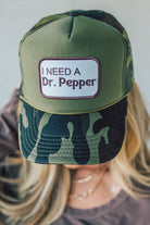 Blonde woman wearing a trucker hat with a patch on front with a patch that says "I Need a Dr. Pepper" and a camo print bill, and mesh back.