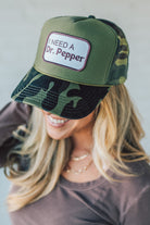 Blonde woman wearing a trucker hat with a patch on front with a patch that says "I Need a Dr. Pepper" and a camo print bill, and mesh back.