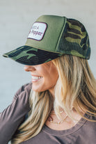 Blonde woman wearing a trucker hat with a patch on front with a patch that says "I Need a Dr. Pepper" and a camo print bill, and mesh back.