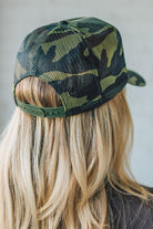 Blonde woman wearing a trucker hat with a patch on front with a patch that says "I Need a Dr. Pepper" and a camo print bill, and mesh back.