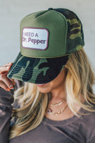 Blonde woman wearing a trucker hat with a patch on front with a patch that says "I Need a Dr. Pepper" and a camo print bill, and mesh back.