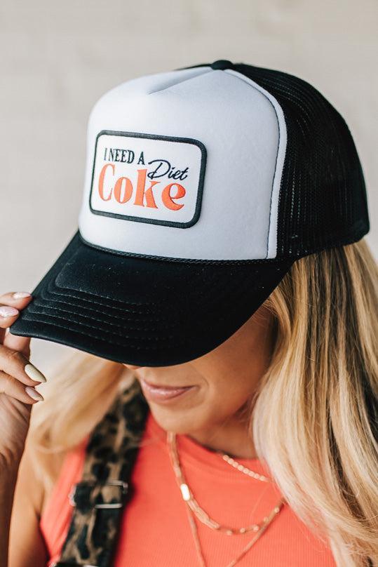 Blonde woman wearing a black and white trucker hat with a "NEED A DIET COKE" patch.