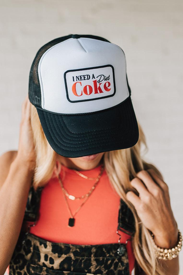 Blonde woman wearing a black and white trucker hat with a "NEED A DIET COKE" patch.