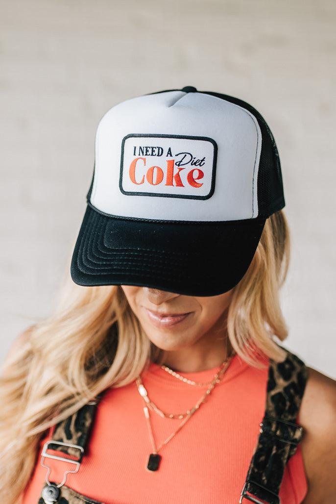 Blonde woman wearing a black and white trucker hat with a "NEED A DIET COKE" patch.
