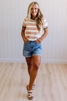 Woman wearing a striped shirt and denim shorts standing against a white wall.