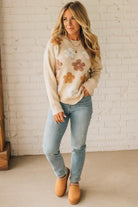 Woman wearing a floral sweater and jeans standing against a white brick wall.