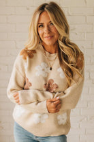 Woman wearing a beige sweater with floral patterns against a white brick wall.