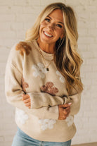 Woman wearing a beige sweater with floral patterns against a white brick wall.
