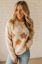 Woman wearing a beige sweater with floral patterns against a white brick wall.