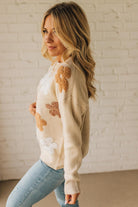Woman wearing a beige sweater with floral patterns against a white brick wall.