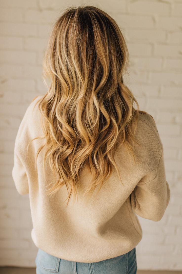 Person wearing a beige sweater with long, wavy hair against a white brick wall.