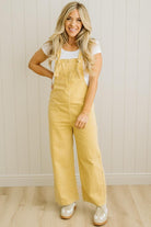 Woman wearing mustard yellow overalls against a white paneled wall.