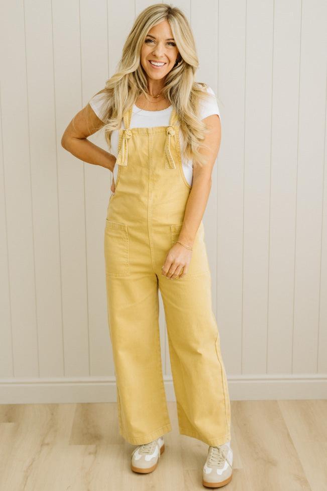 Woman wearing mustard yellow overalls against a white paneled wall.