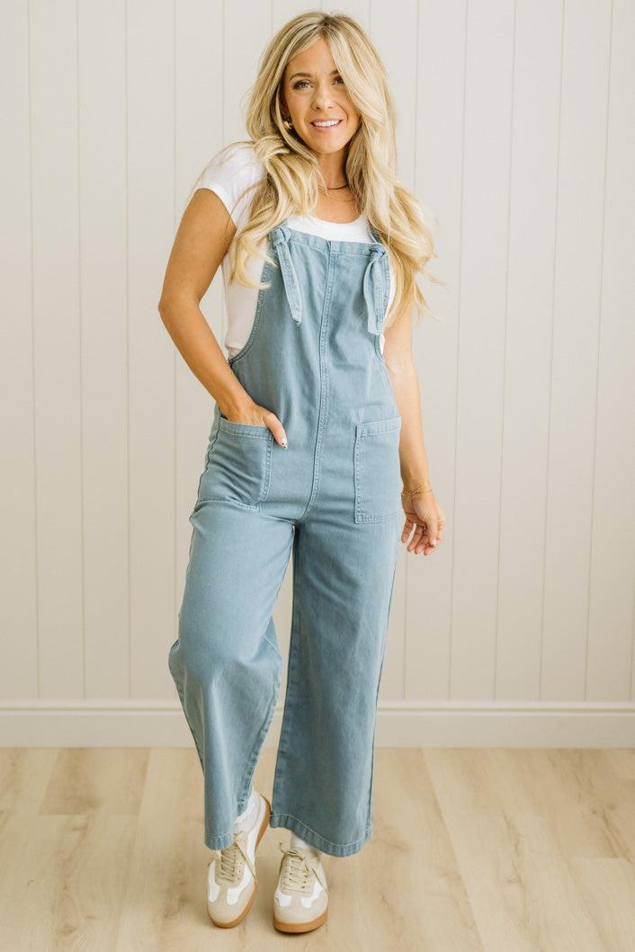 Woman wearing light blue overalls against a white paneled wall.