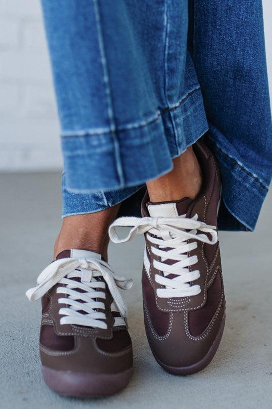 Brown sneakers with white laces worn with blue jeans on a light gray floor.