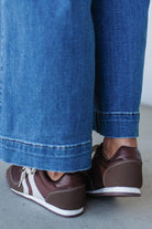 Brown shoes with white soles worn with blue jeans on a light gray floor.