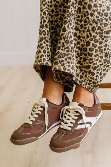 Brown sneakers with white laces worn with leopard jeans on a light wood floor.