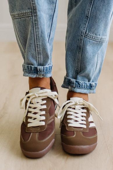 Brown sneakers with white laces worn with leopard jeans on a light wood floor.