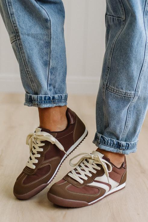 Brown sneakers with white laces worn with leopard jeans on a light wood floor.