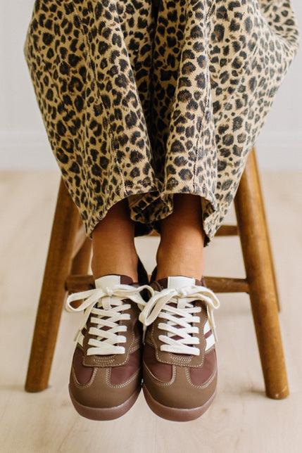 Brown sneakers with white laces worn with leopard jeans on a light wood floor.