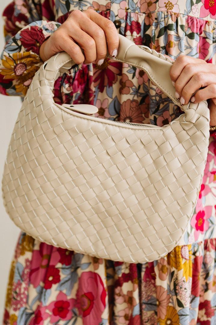 Beige woven handbag held by a person wearing a floral dress.