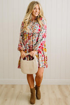 Woman wearing a floral dress holding a beige handbag against a white paneled wall.