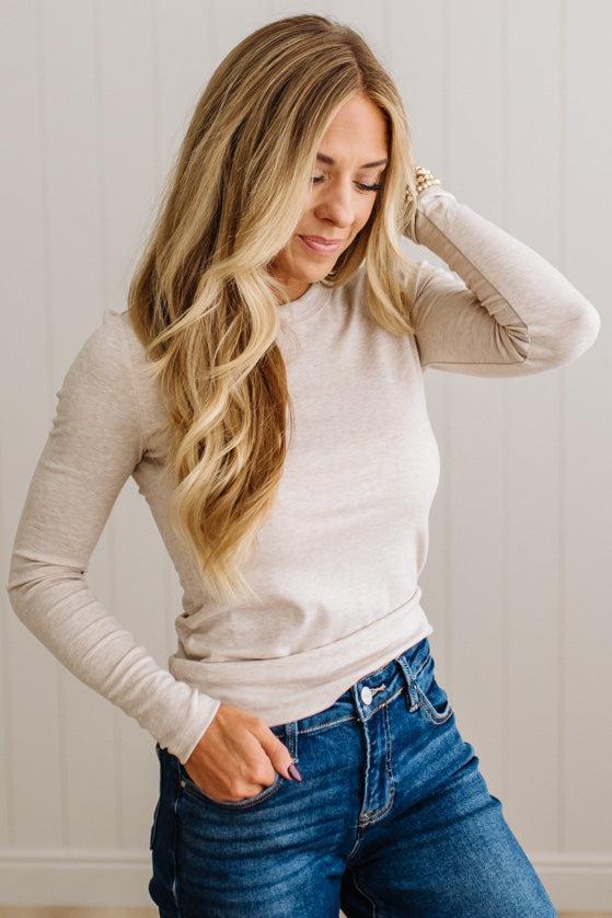 A woman with long blonde hair wearing a beige long-sleeve knit top and blue jeans, posing against a white paneled wall.