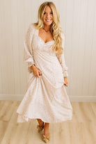 Woman wearing a white floral dress standing against a light wooden paneled wall.