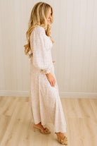 Woman wearing a white floral dress standing against a light wooden paneled wall.