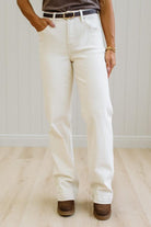 A person wearing off-white straight-leg jeans with a black belt, paired with brown suede boots, standing in a room with light wood floors and white paneled walls.