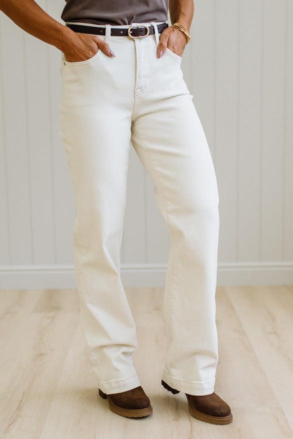 A person wearing off-white or cream-colored straight-leg jeans, styled with a dark belt, a taupe top, and brown suede boots, standing on a light wood floor against a white paneled wall.