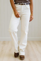 A person wearing off-white straight-leg jeans with a dark belt, paired with brown boots, standing on a light wood floor against a white paneled wall.
