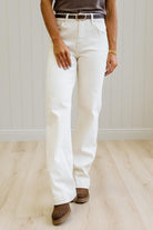 A person wearing off-white straight-leg jeans with a brown belt and brown suede boots, standing on a light wood floor against a white paneled wall.