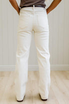 A person shown from the back wearing off-white or cream-colored straight-leg jeans, paired with a dark brown top and dark brown shoes, standing on a light wood floor against a white paneled wall.