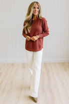 A woman modeling a rust-colored long-sleeve knit top and off-white wide-leg pants in a bright room with light wood floors and white paneled walls.