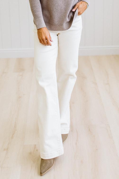 A person wearing white wide-leg jeans, a taupe knit sweater, and taupe pointed-toe flats, standing on a light wood floor against a white paneled wall.