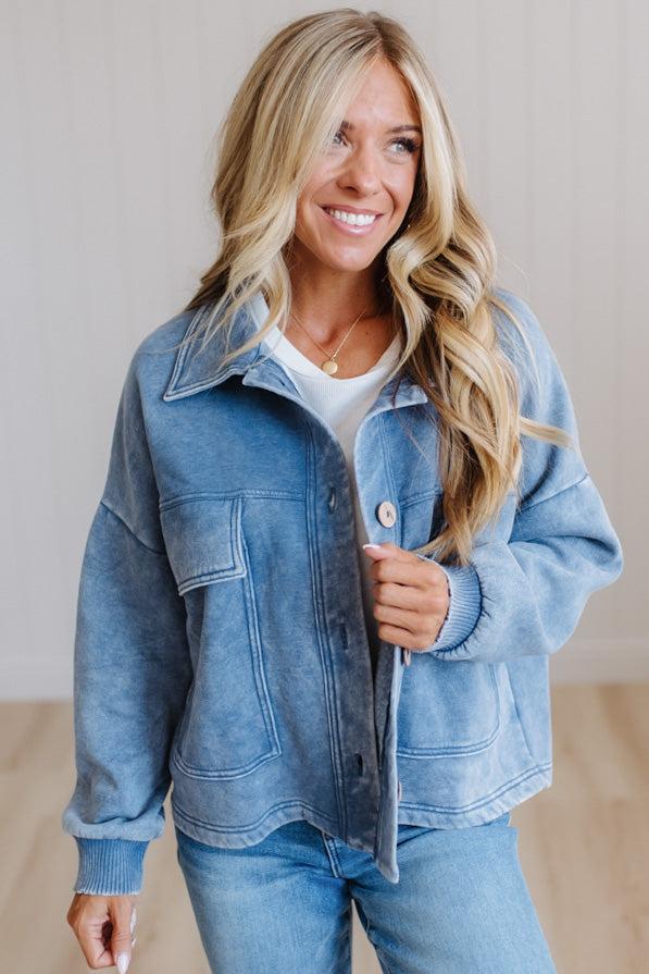A smiling blonde woman wearing a light blue washed denim jacket over a white top and blue jeans, standing in a bright indoor setting with a light neutral wall and wood floor.
