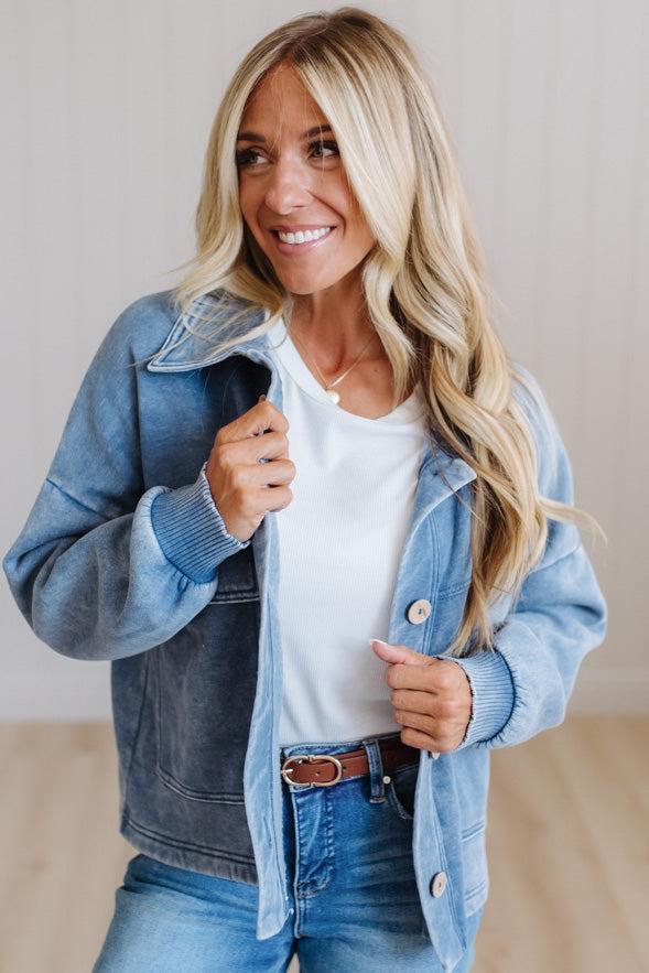 A smiling blonde woman wearing a light blue denim jacket over a white t-shirt and blue jeans, standing against a light beige wall.