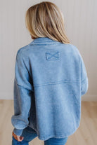 A person viewed from behind wearing a light blue washed denim jacket with a small rectangular patch detail on the upper back, paired with blue jeans in a bright indoor setting.