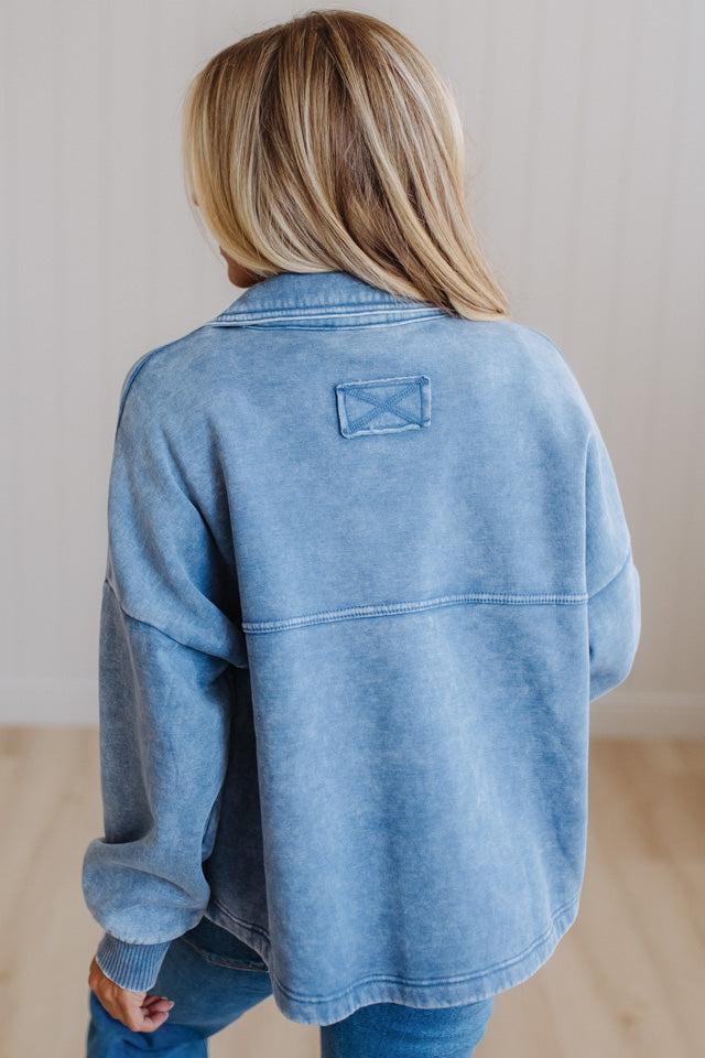 A person viewed from behind wearing a light blue washed denim jacket with a small rectangular patch detail on the upper back, paired with blue jeans in a bright indoor setting.