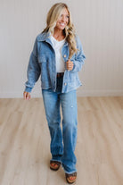A woman modeling a light blue denim jacket and matching wide-leg jeans, styled with a white t-shirt, dark brown belt, and brown slide sandals, standing on a light wood floor against a white paneled wall.