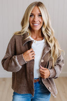 A smiling blonde woman wearing a brown corduroy-style jacket over a white top and blue jeans, standing indoors against a light neutral wall.