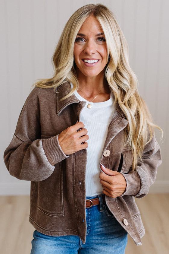 A smiling blonde woman wearing a brown corduroy-style jacket over a white top and blue jeans, standing indoors against a light neutral wall.