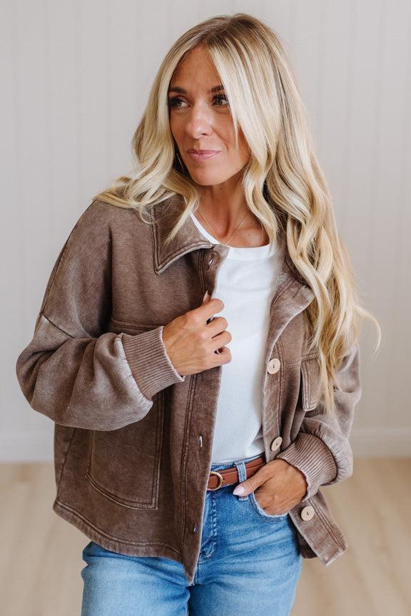 A woman wearing a brown suede-like button-front jacket over a white top and blue jeans, standing indoors against a light neutral wall.