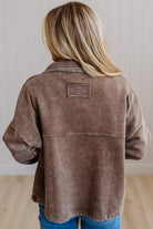 A woman viewed from behind wearing a brown acid-wash button-up shirt jacket with a small rectangular patch on the back, paired with blue jeans.