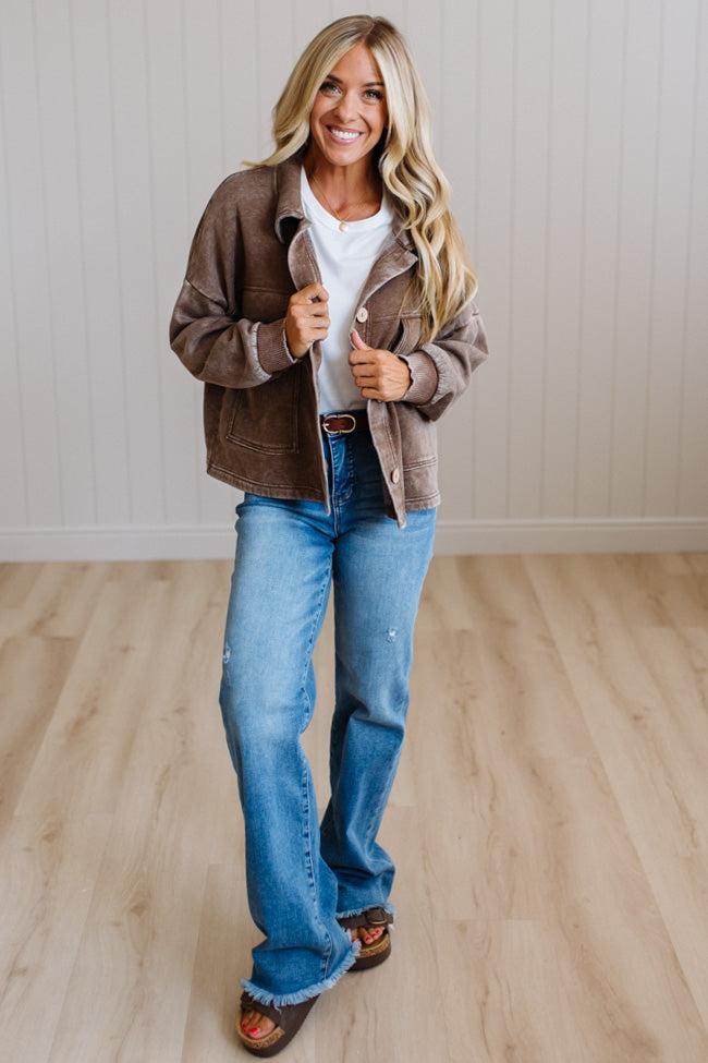 A woman modeling a brown suede jacket over a white t-shirt and blue jeans, standing in a room with light wood floors and white paneled walls.