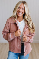 A smiling blonde woman wearing a dusty rose pink corduroy jacket over a white t-shirt and blue jeans, standing in a brightly lit indoor setting.
