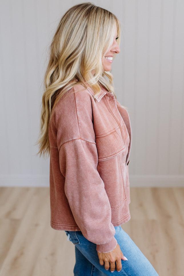 A woman wearing a dusty rose pink cropped button-up jacket with a soft, textured fabric, paired with blue jeans, shown from a side profile against a light wood-paneled wall.