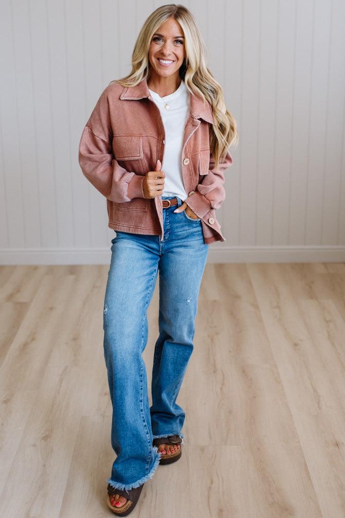 A woman modeling a dusty rose utility jacket over a white t-shirt and blue flare jeans, standing on a light wood floor against a white paneled wall.