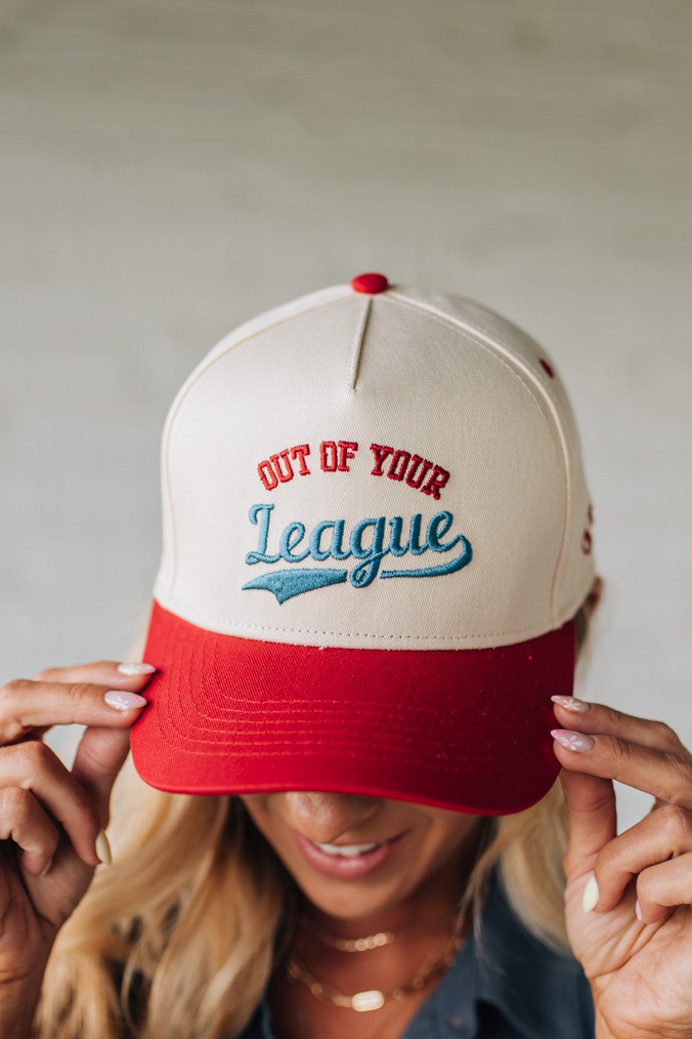 Blonde woman wearing a trucker hat in cream with red brim, and red and blue embroidery at the front "Out of Your League"